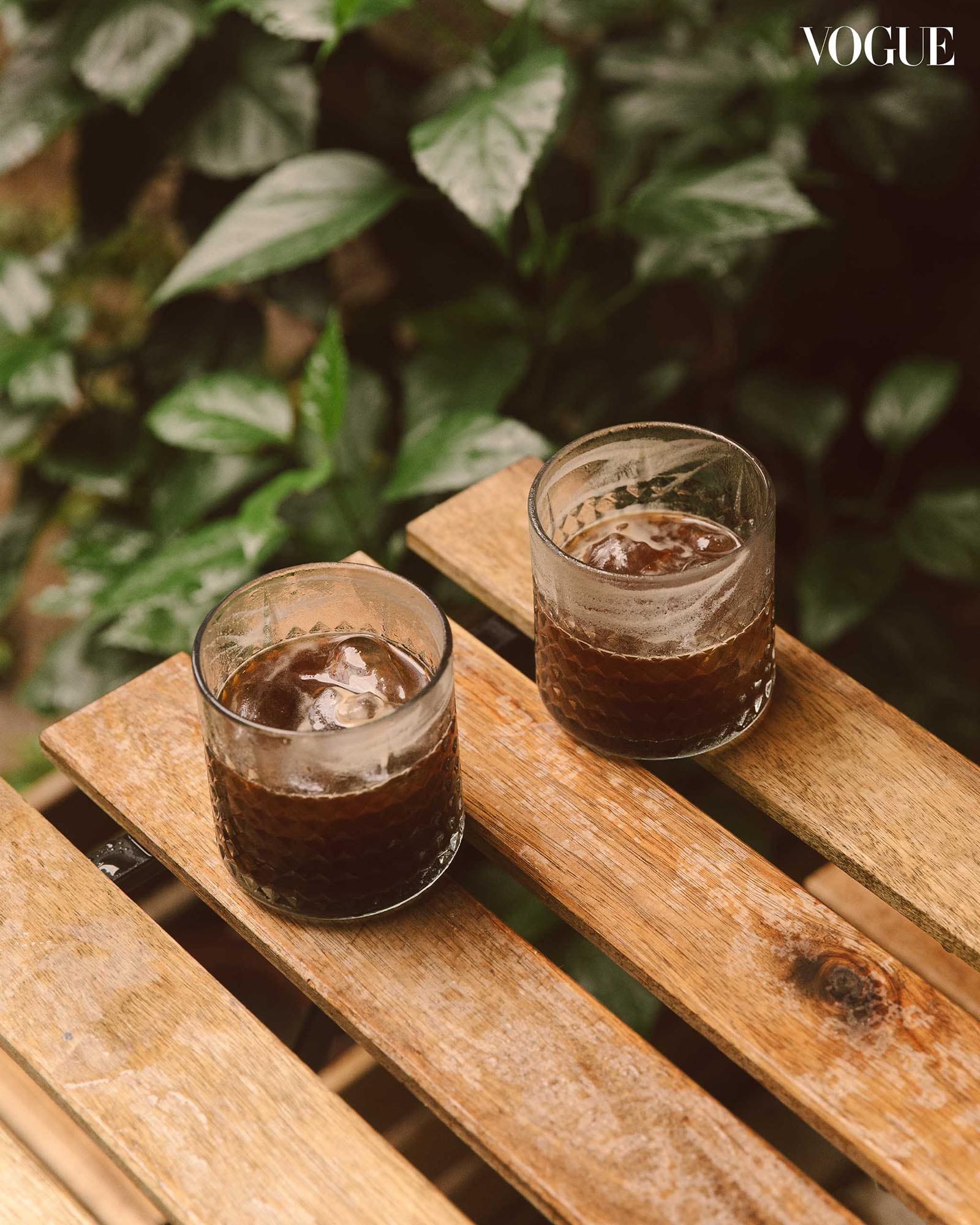 Two glasses of iced coffee on a wooden pallet table surrounded by lush greenery